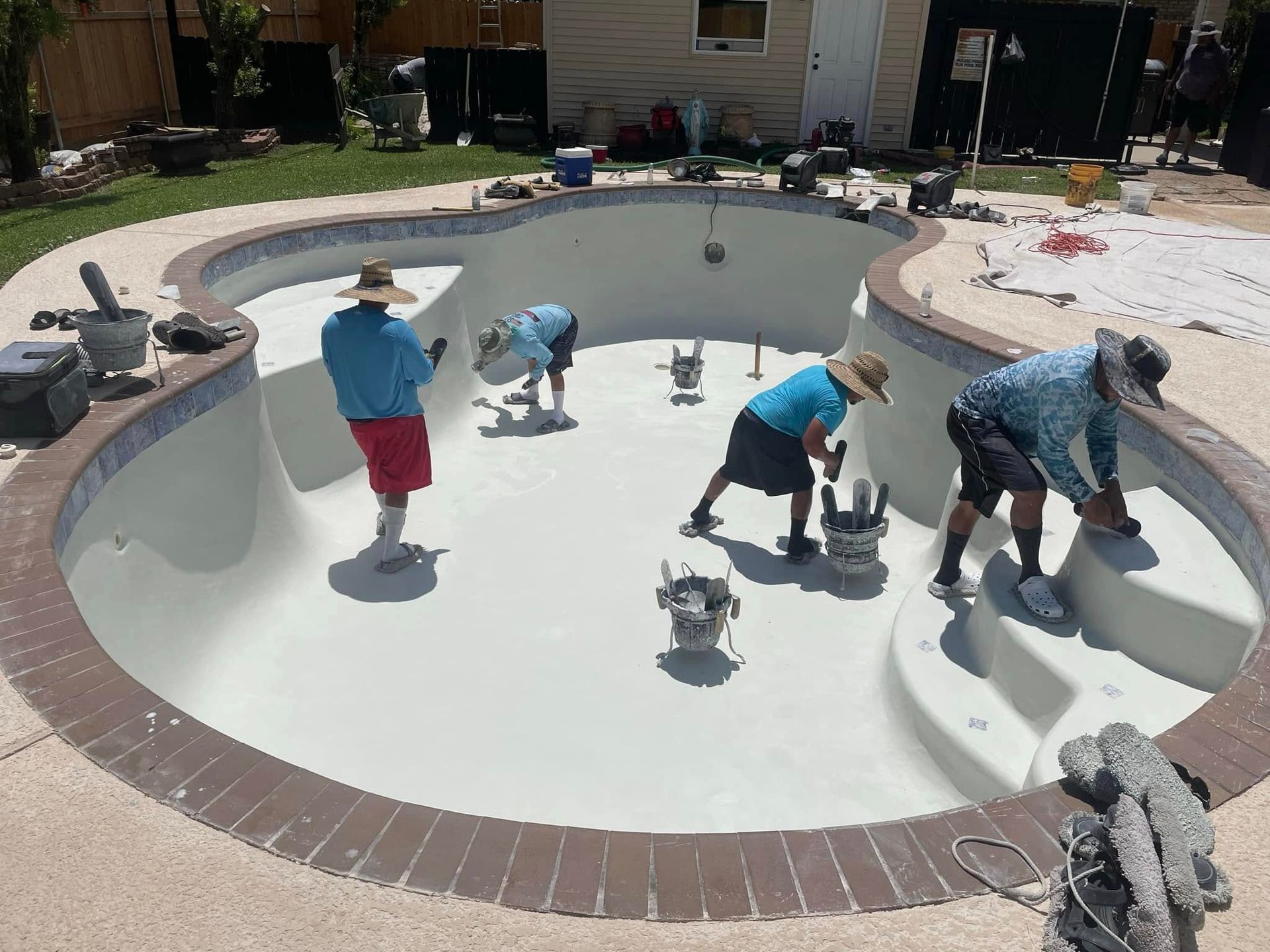 Children cleaning an empty backyard pool with brushes and buckets on a sunny day