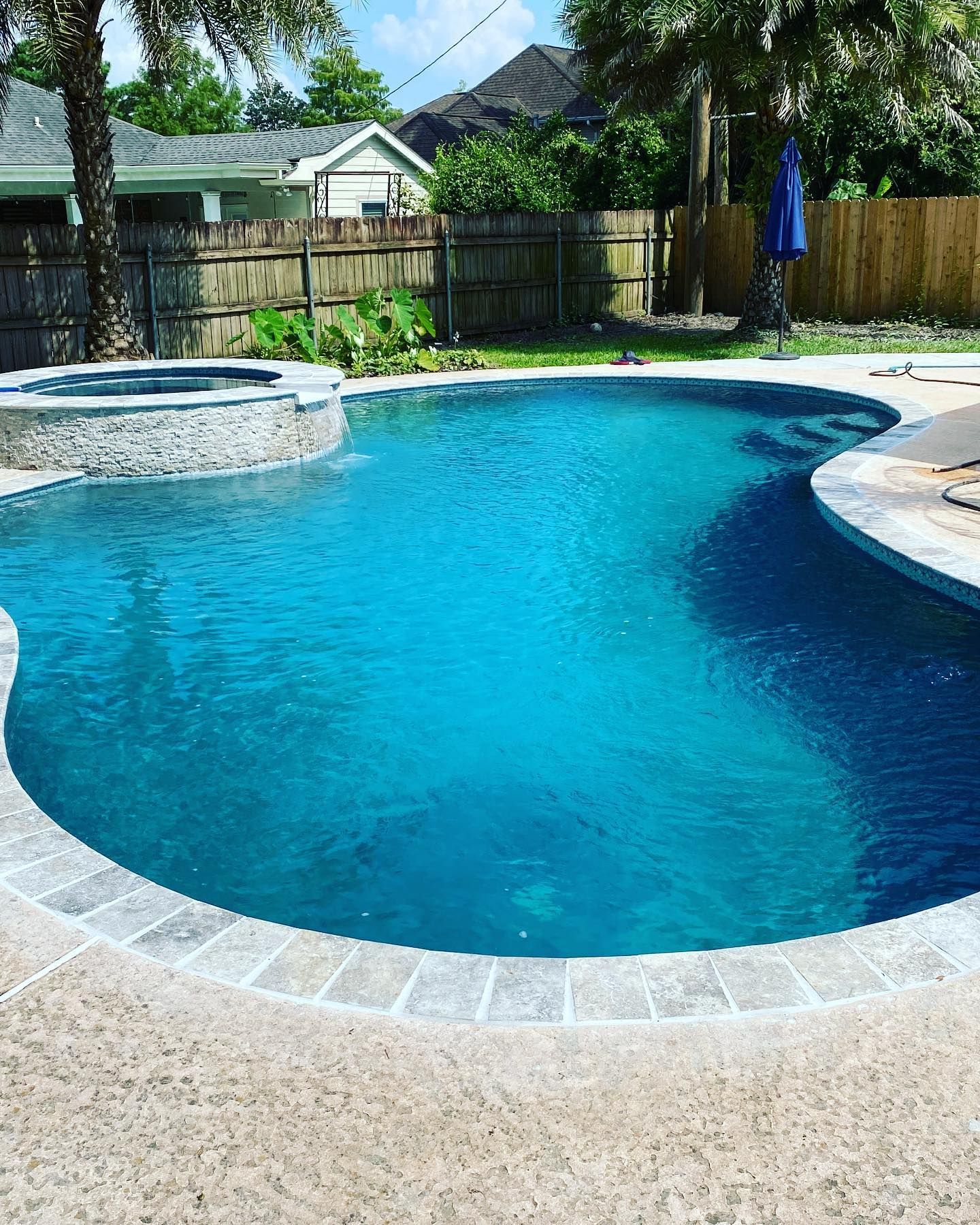 Backyard swimming pool with blue water, stone edge, and a wooden fence in the background