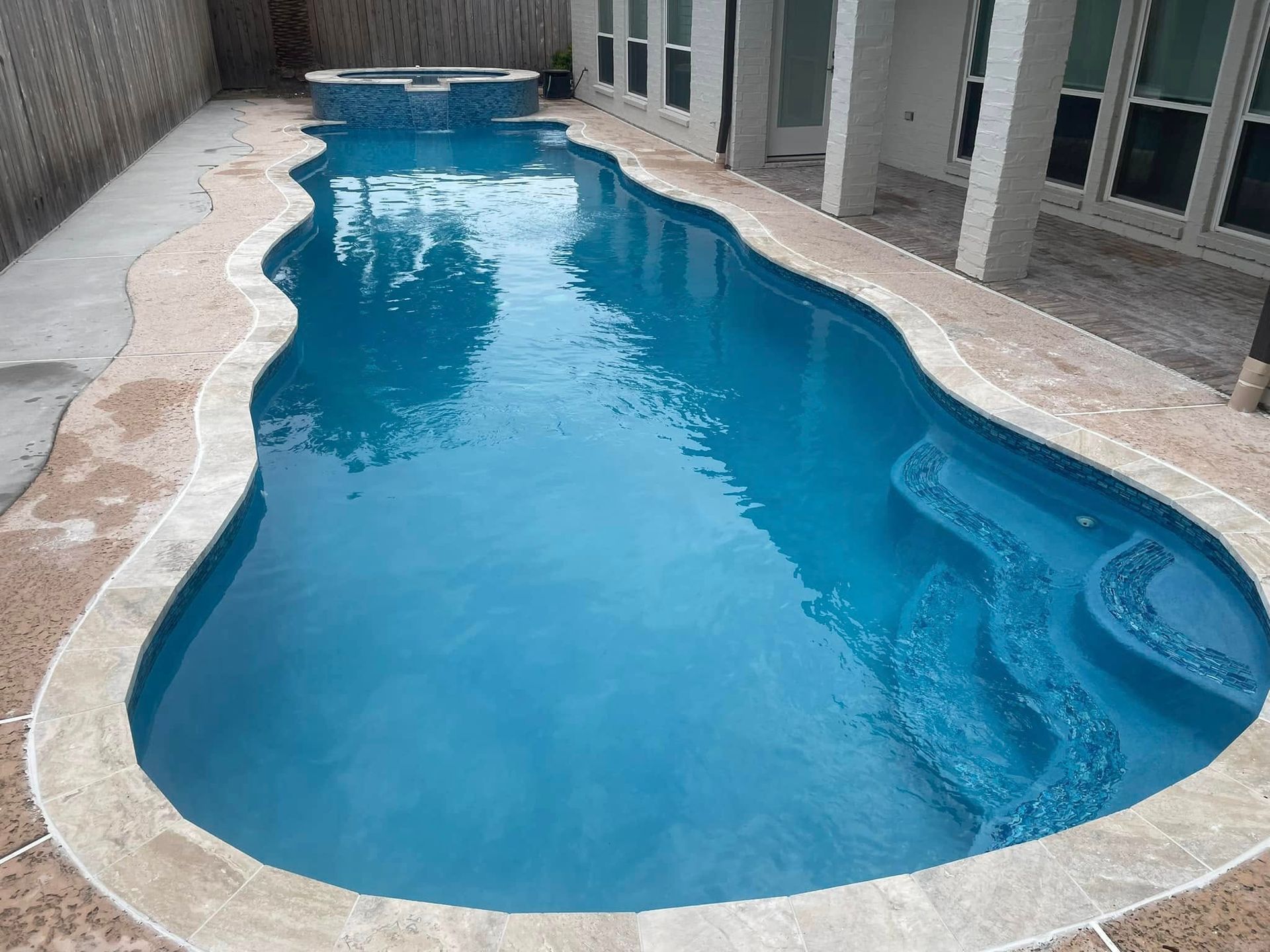 Narrow blue swimming pool beside a house, with tan stone decking and built-in steps on one end