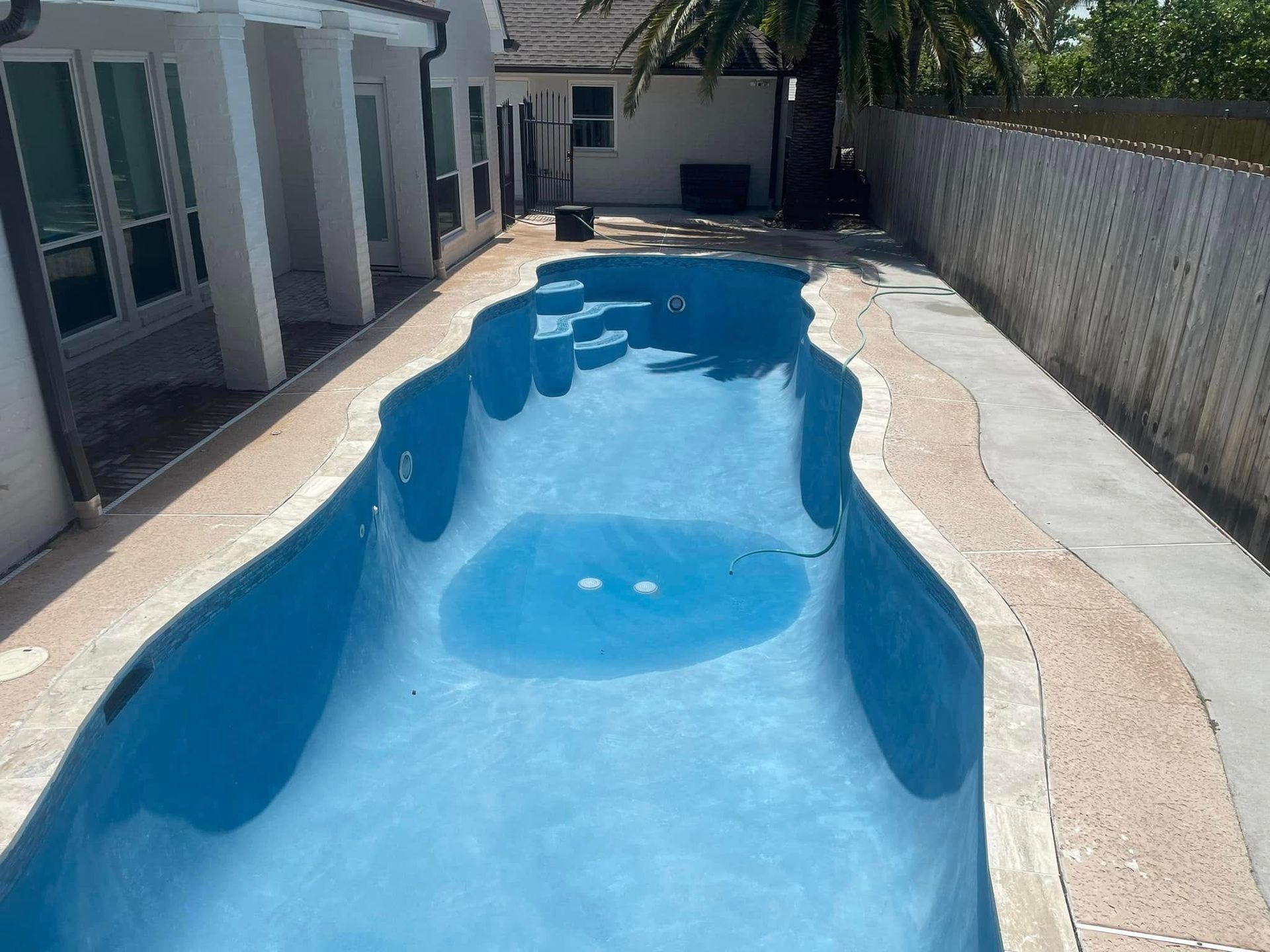 Narrow blue swimming pool beside a white house and fence in a sunny backyard.
