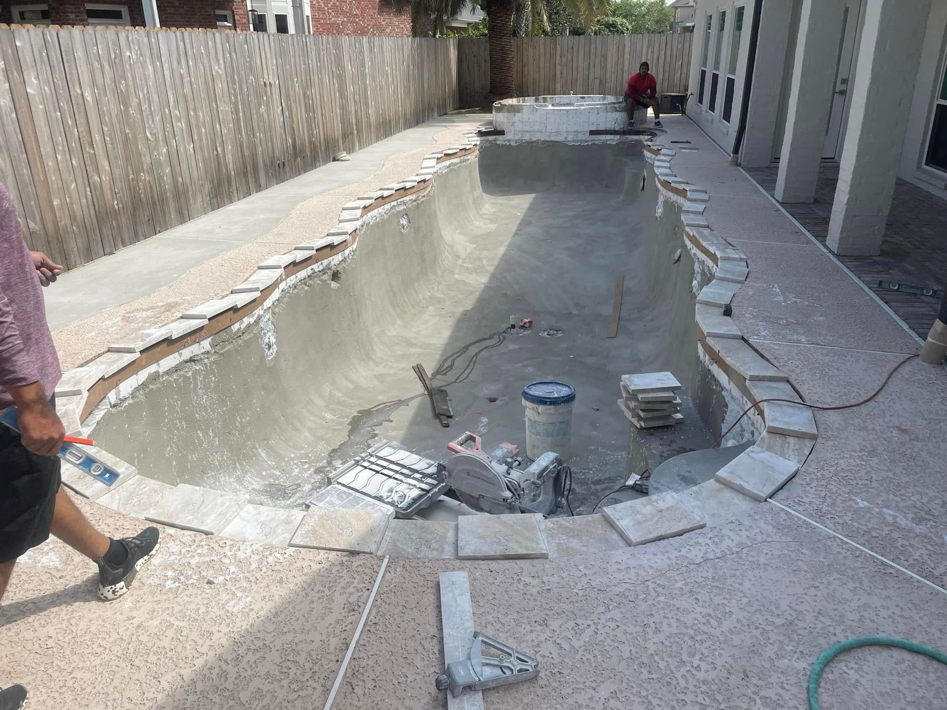 Outdoor swimming pool under renovation, with workers, broken tiles, and tools along the deck.