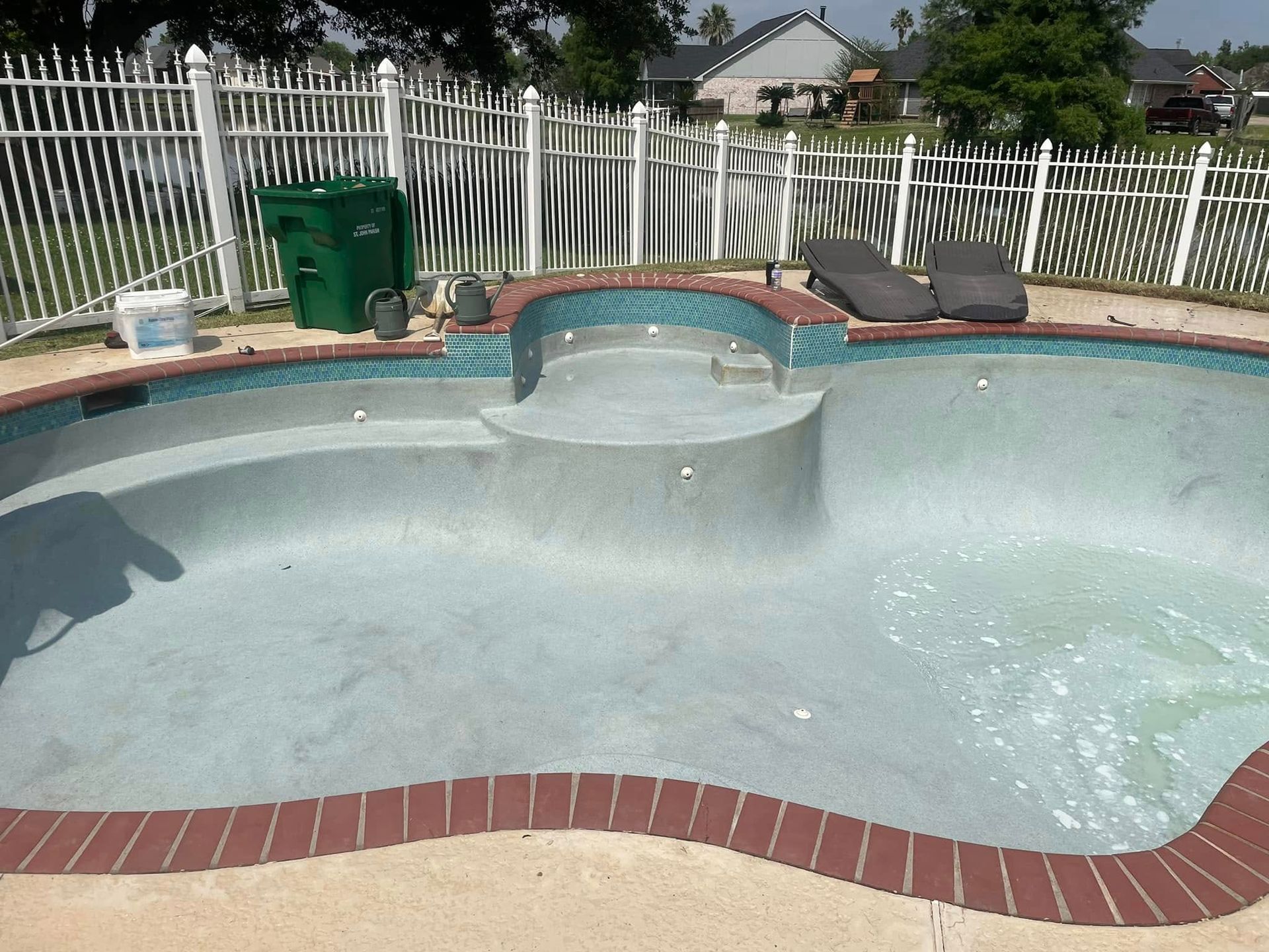 Empty backyard swimming pool with a damaged, peeling interior and brick coping, near a white fence.