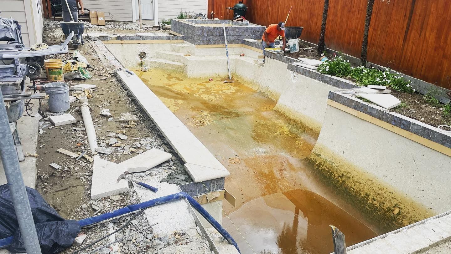 Construction site with a rectangular concrete pool filled with muddy water and surrounded by unfinished pathways.