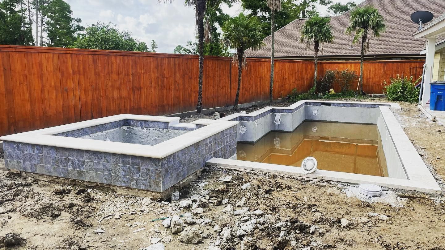 Rectangular concrete pool under construction beside a wooden fence, with muddy ground and trees nearby