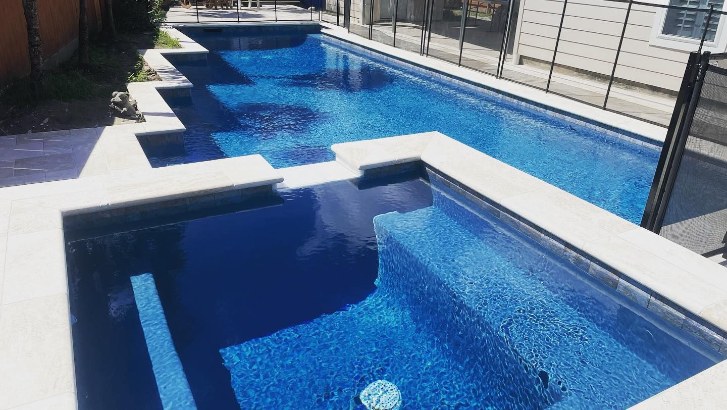 Blue swimming pool with shallow and deep sections beside a walkway and fence in bright sunlight