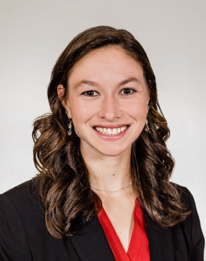 Woman smiling, wearing a black blazer over a red top. Dark, wavy hair. White background.