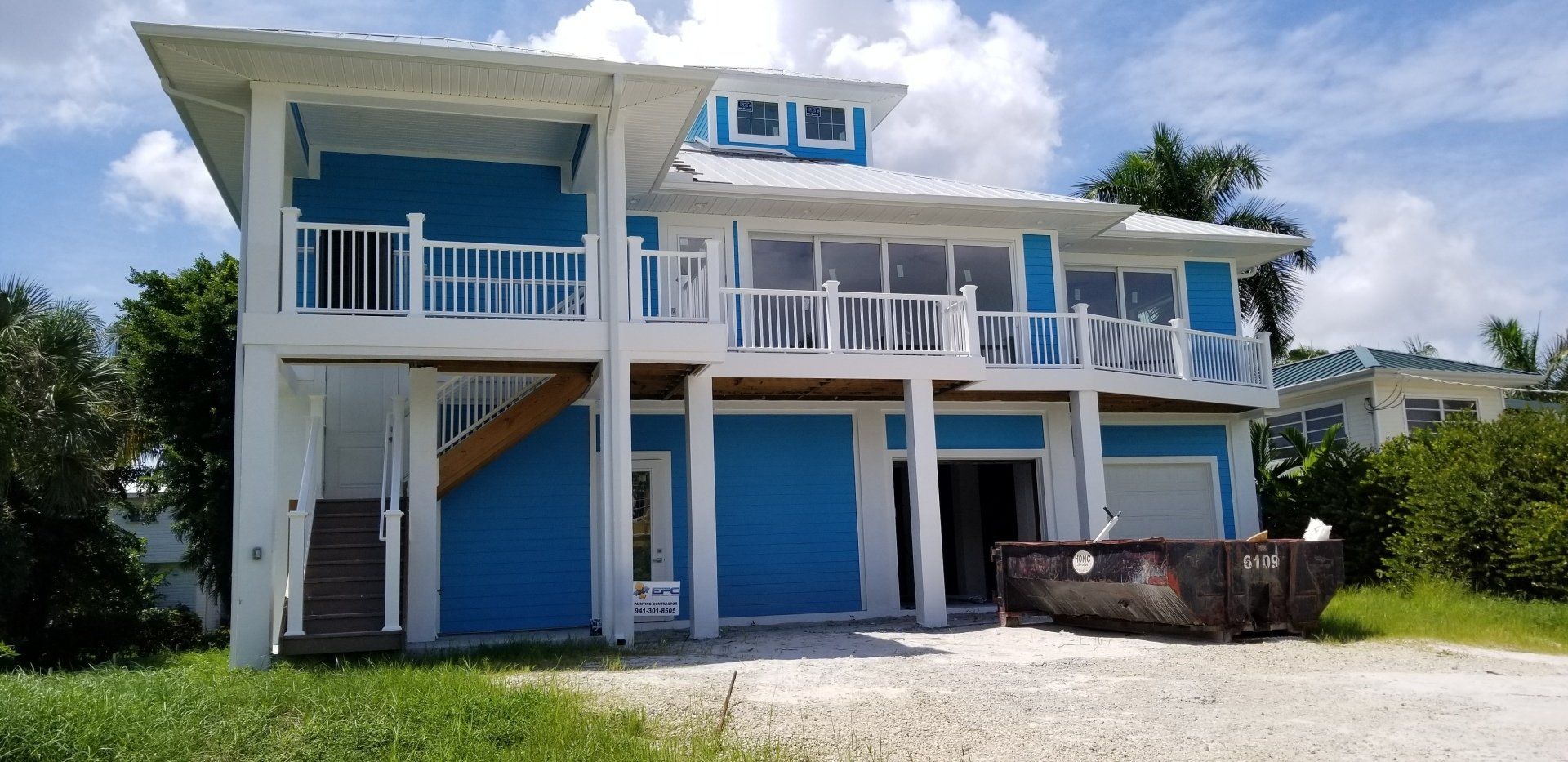 Blue and white house with a balcony and a dumpster in the driveway.