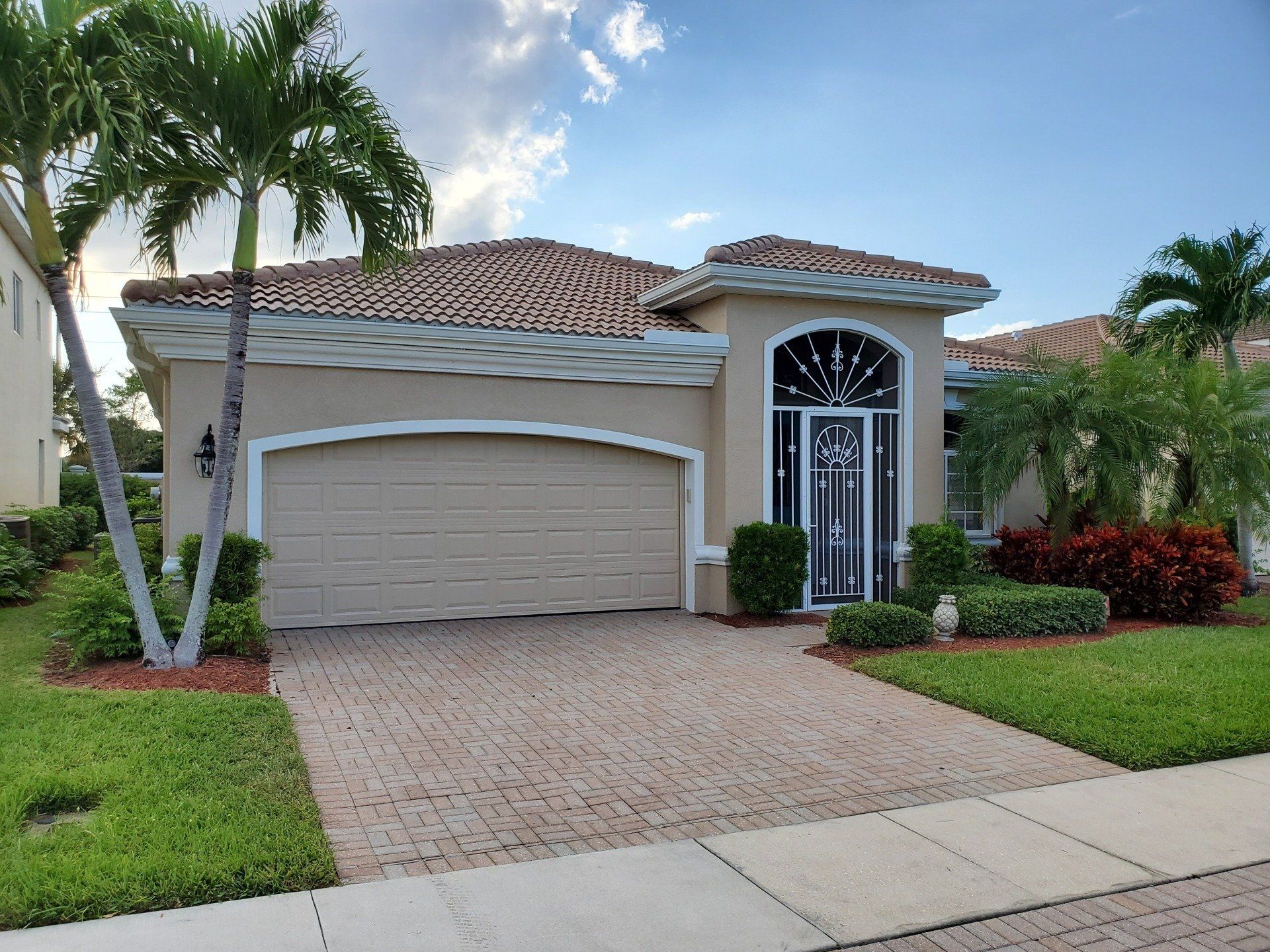 Tan house with a brick driveway, palm trees, and a beige garage door.