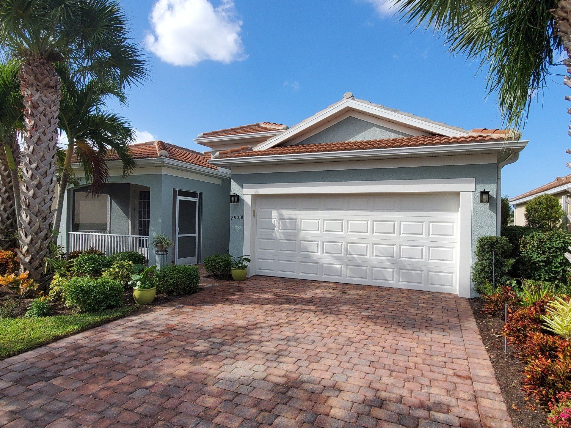 Blue house with red brick driveway, white garage door, and palm trees under a blue sky.