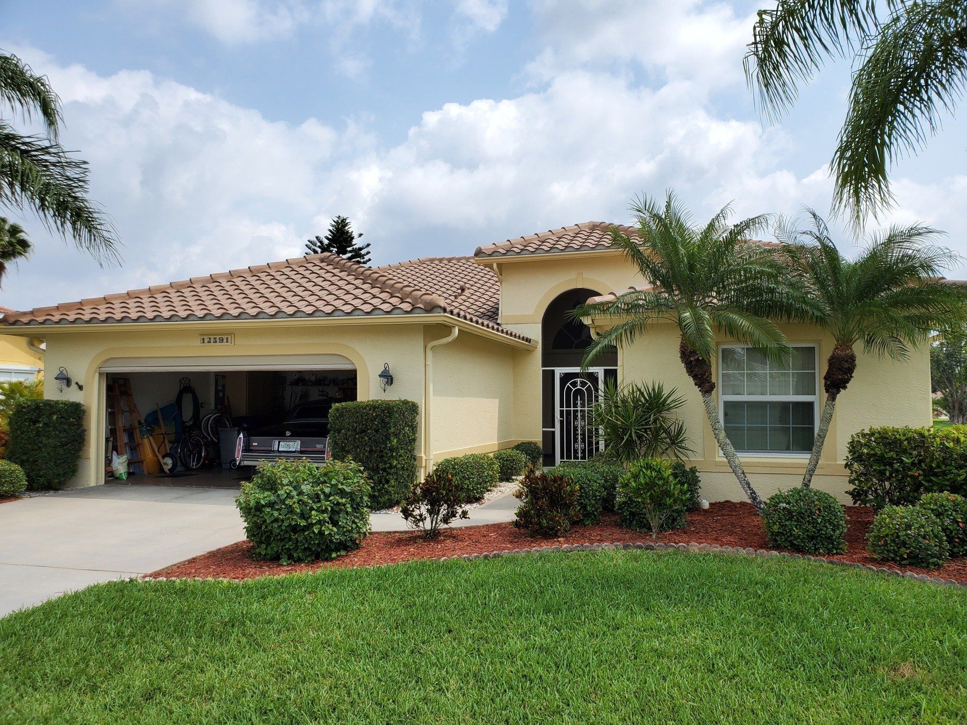 Beige house with red tile roof, garage, green bushes, palm trees, and cloudy sky.