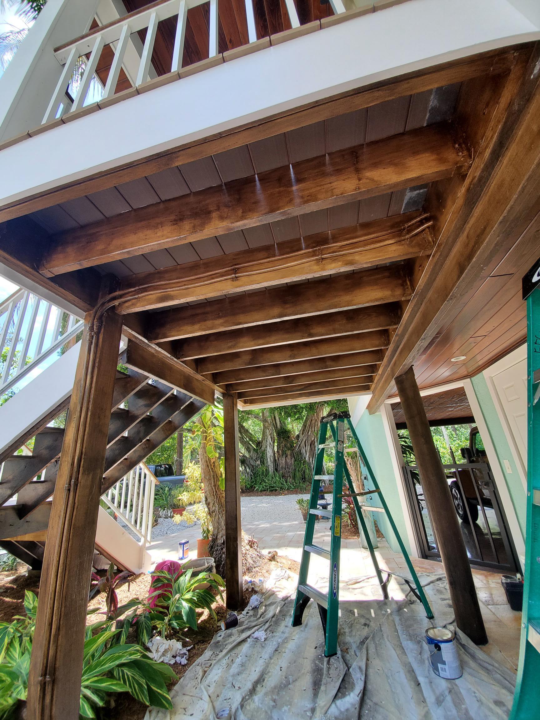 Wooden deck being stained, with stairs, a ladder, and some foliage below.