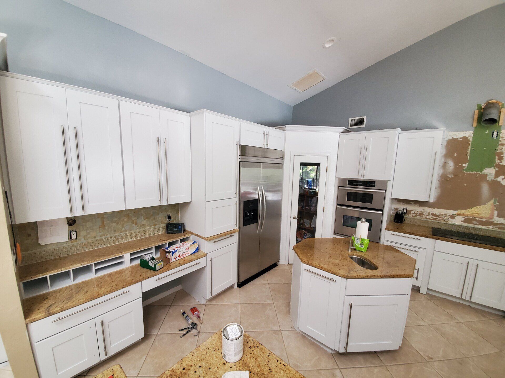 White kitchen cabinets, stainless steel fridge, island, and granite countertops. Construction in progress.