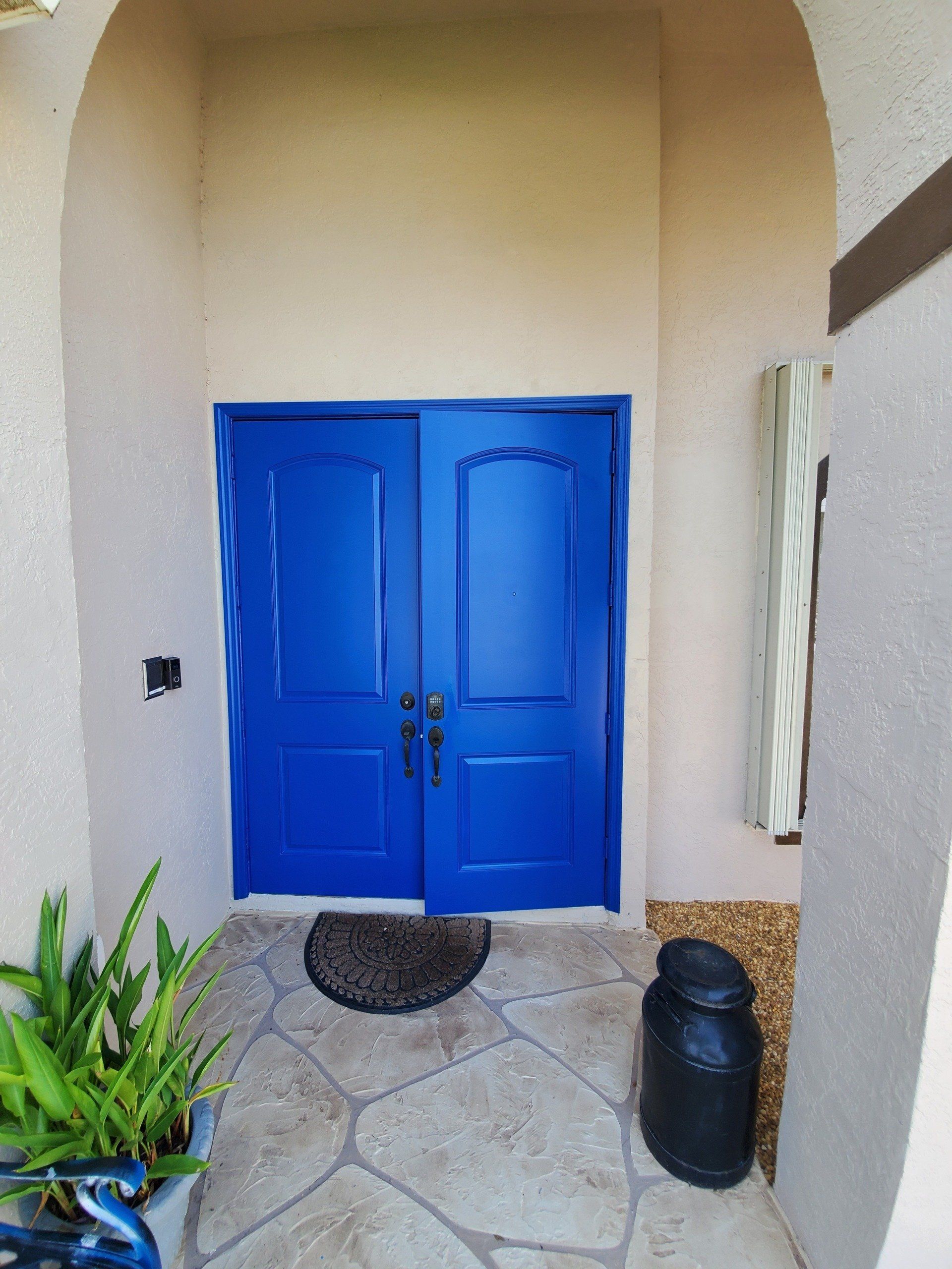 Blue double doors in a beige arched entryway. Slate stone flooring with potted plant on the left and decorative jug on the right.