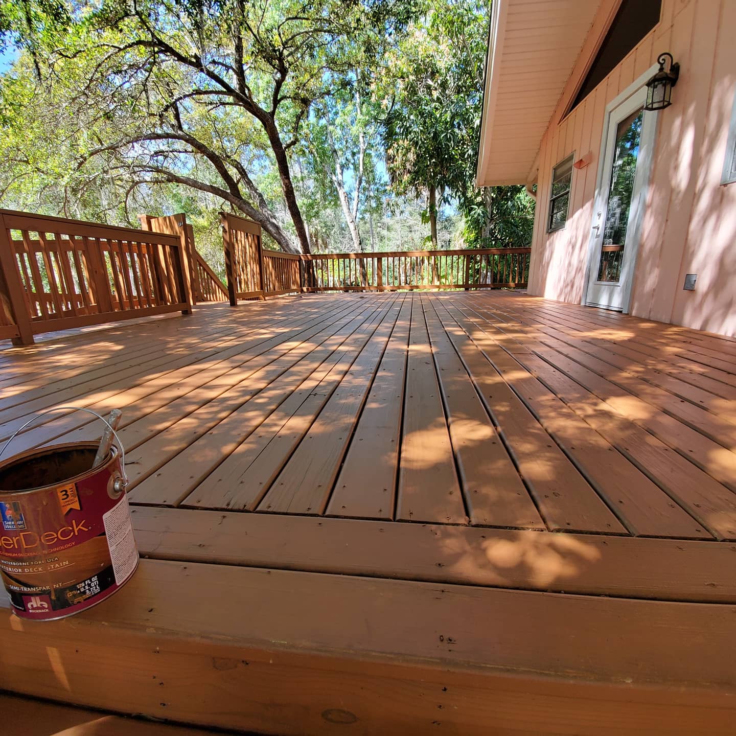 Wooden deck stained brown next to a pink house with open doors, trees in the background.