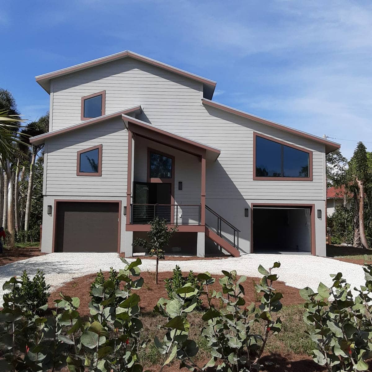 Two-story grey house with brown accents, two garage doors, and slanted windows on a gravel driveway.
