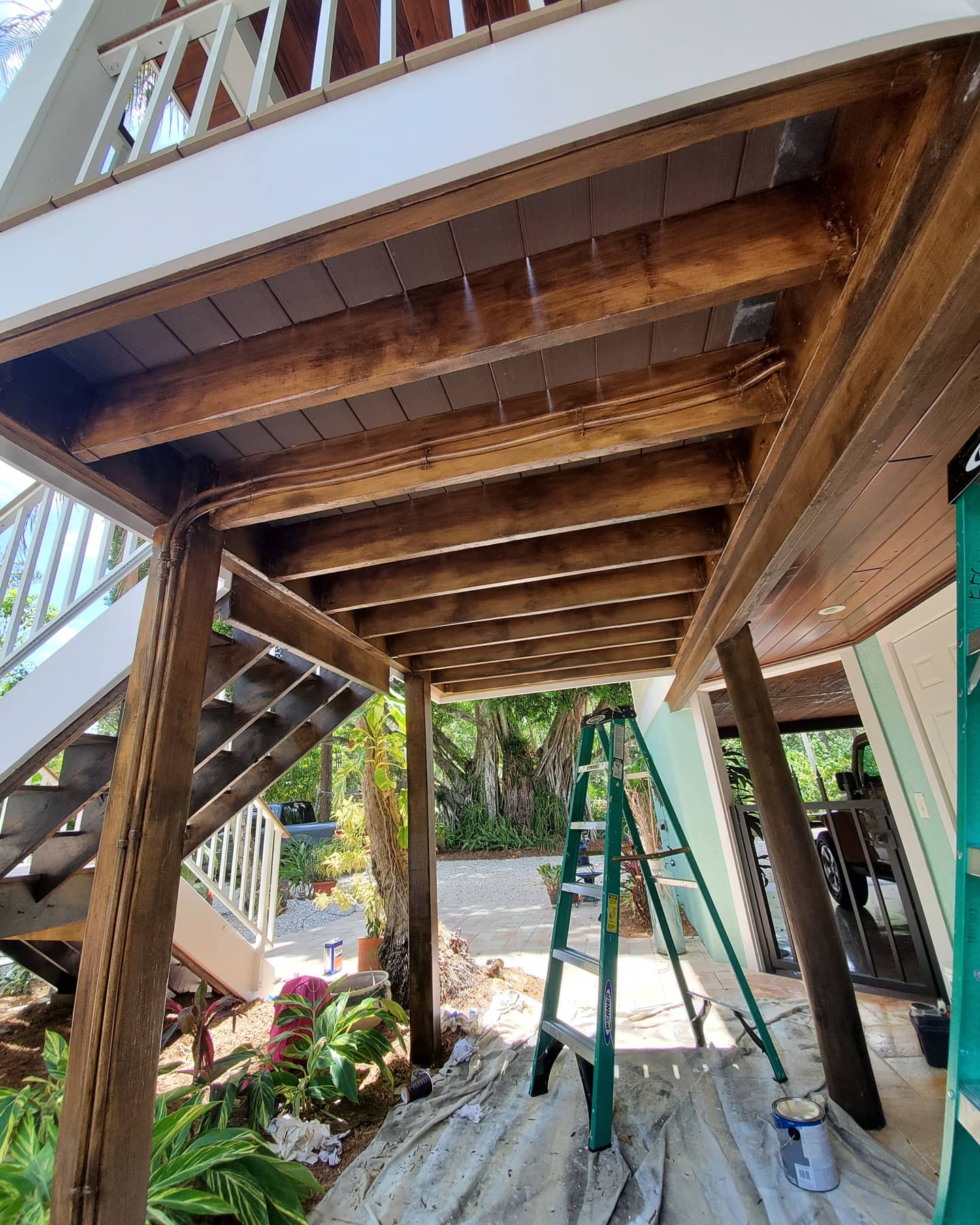 Exterior view of a deck being stained brown with a green ladder and paint cans visible below.