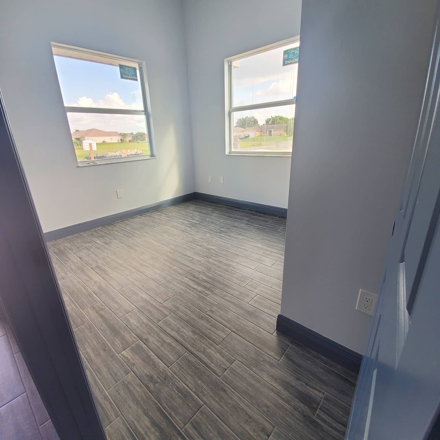 Empty room with gray wood-look flooring, light blue walls, and two windows overlooking a sunny landscape.
