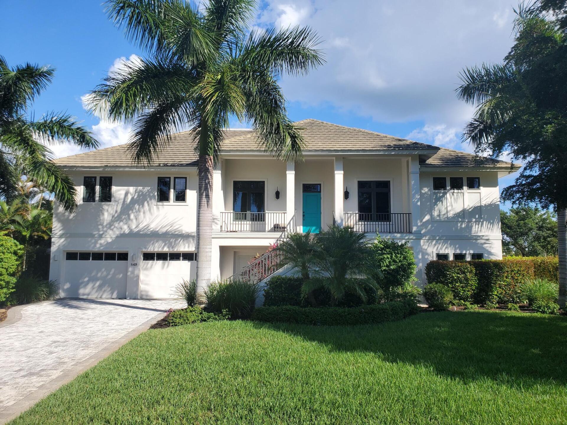 Two-story white house with blue door, palm trees, and a green lawn under a blue sky.