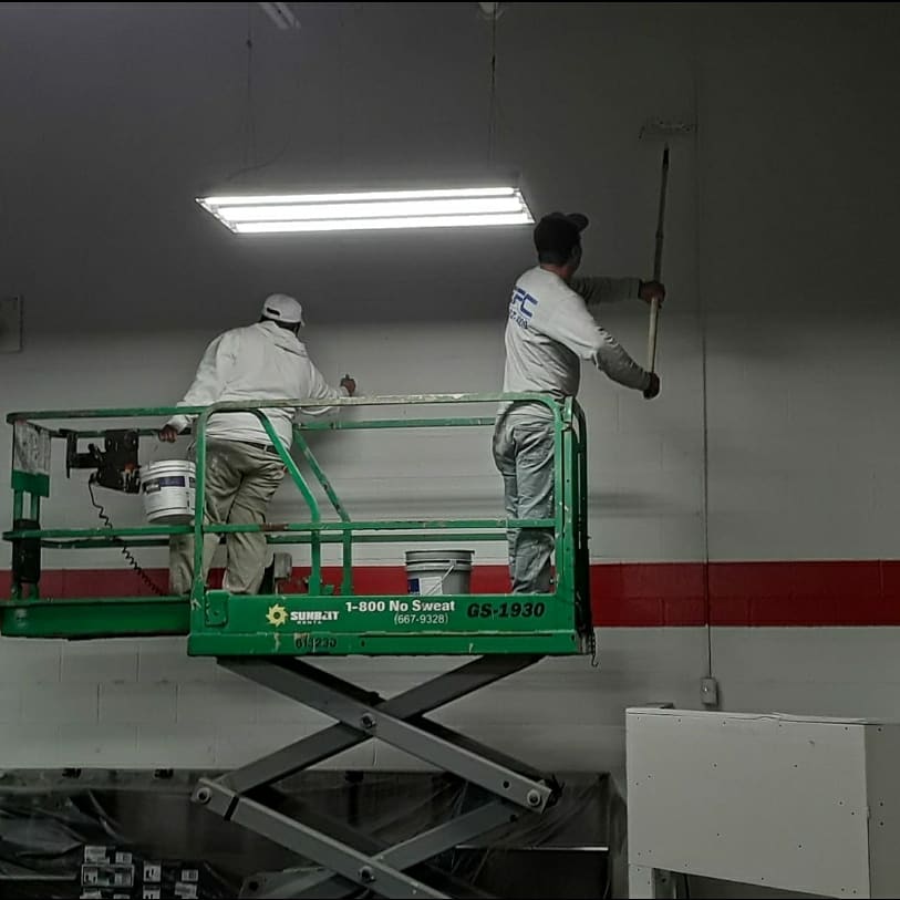 Two painters on a green lift painting a white ceiling in a large room.