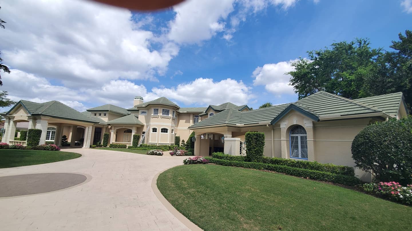 Large beige mansion with green tile roof, circular driveway, and manicured lawn under a blue sky with clouds.