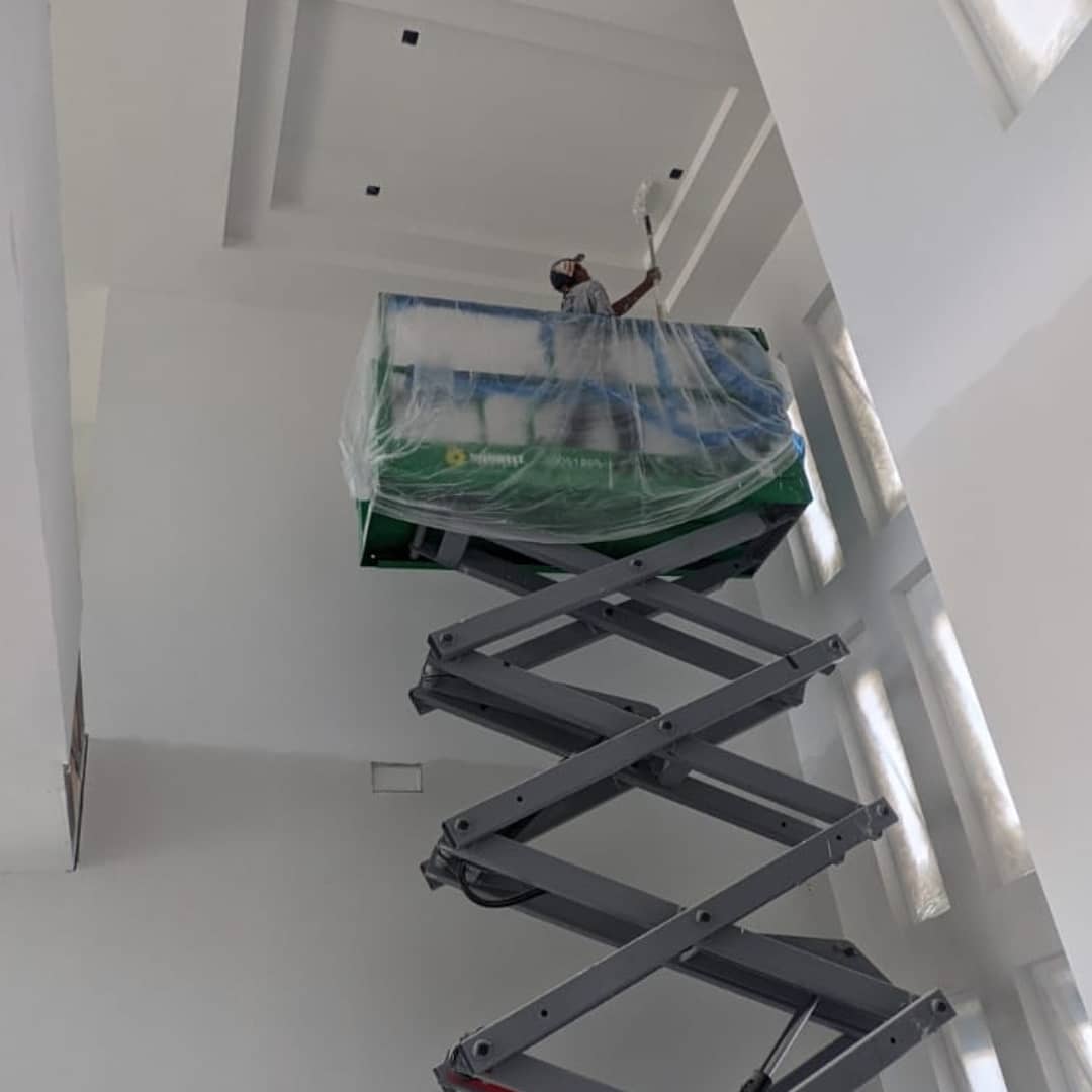 Person painting a high ceiling from a scissor lift. Interior with white walls and natural light.