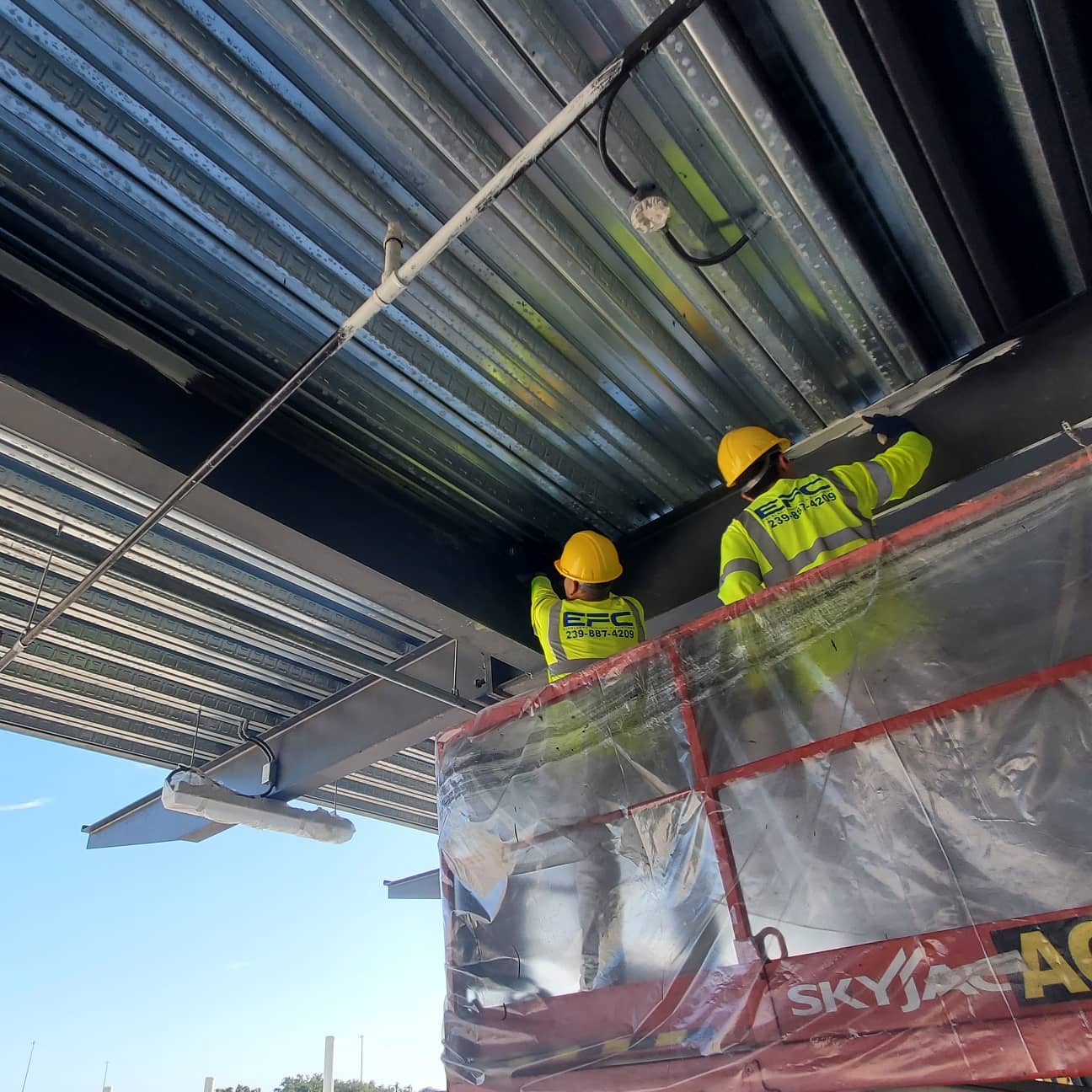 Two construction workers on a lift, wearing yellow hard hats and vests, working on a metal ceiling under a blue sky.