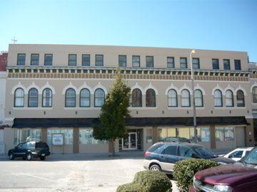 Three-story beige building with arched windows and a central tree on a sunny day with parked cars in front.
