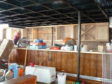 Basement bar under construction: bare wood cabinets against brick wall, dark ceiling, cluttered bar top, person in background.