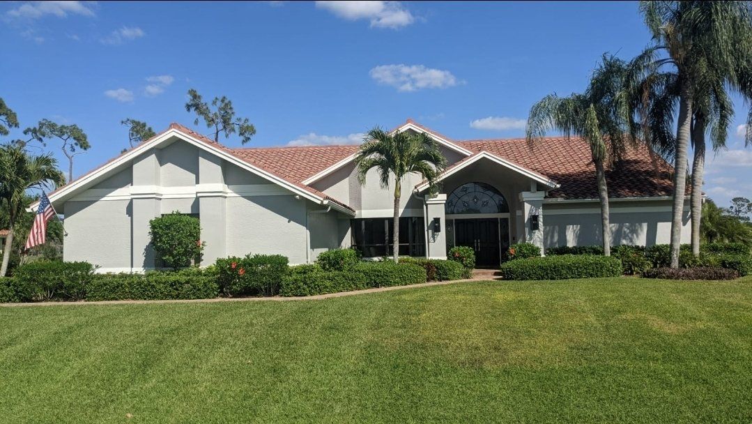 House with red-tiled roof, surrounded by green lawn and tropical foliage under a blue sky.