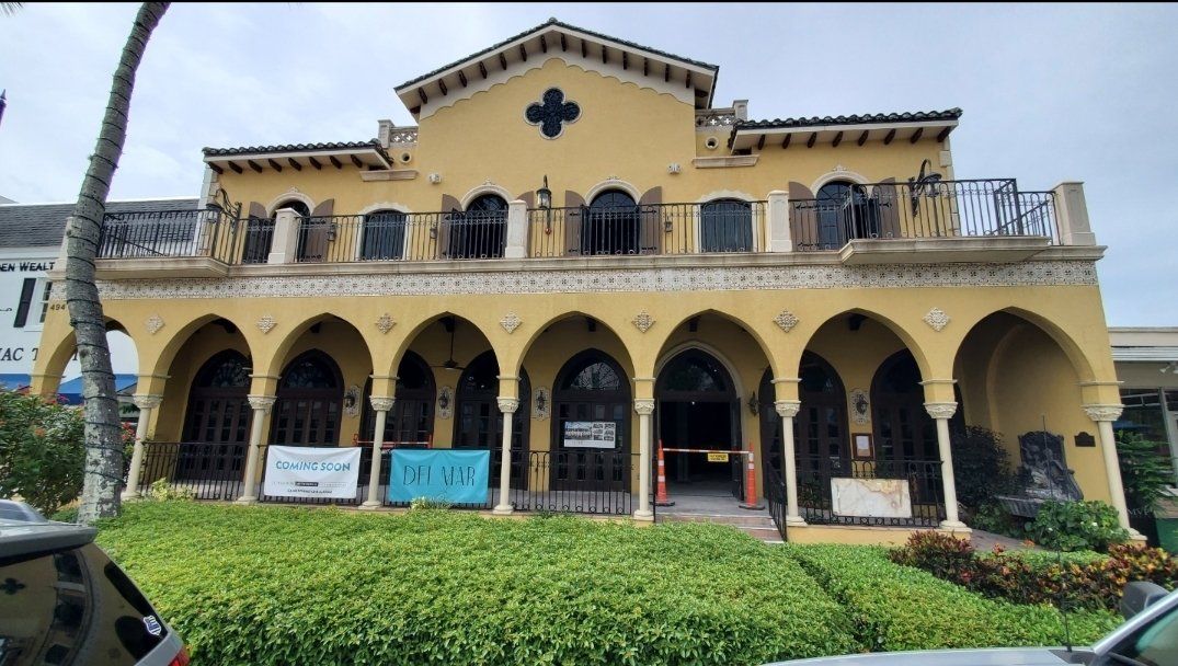 Yellow building with arched facade; street level, two stories. Black iron gates cover the entrances. Green shrubs below.
