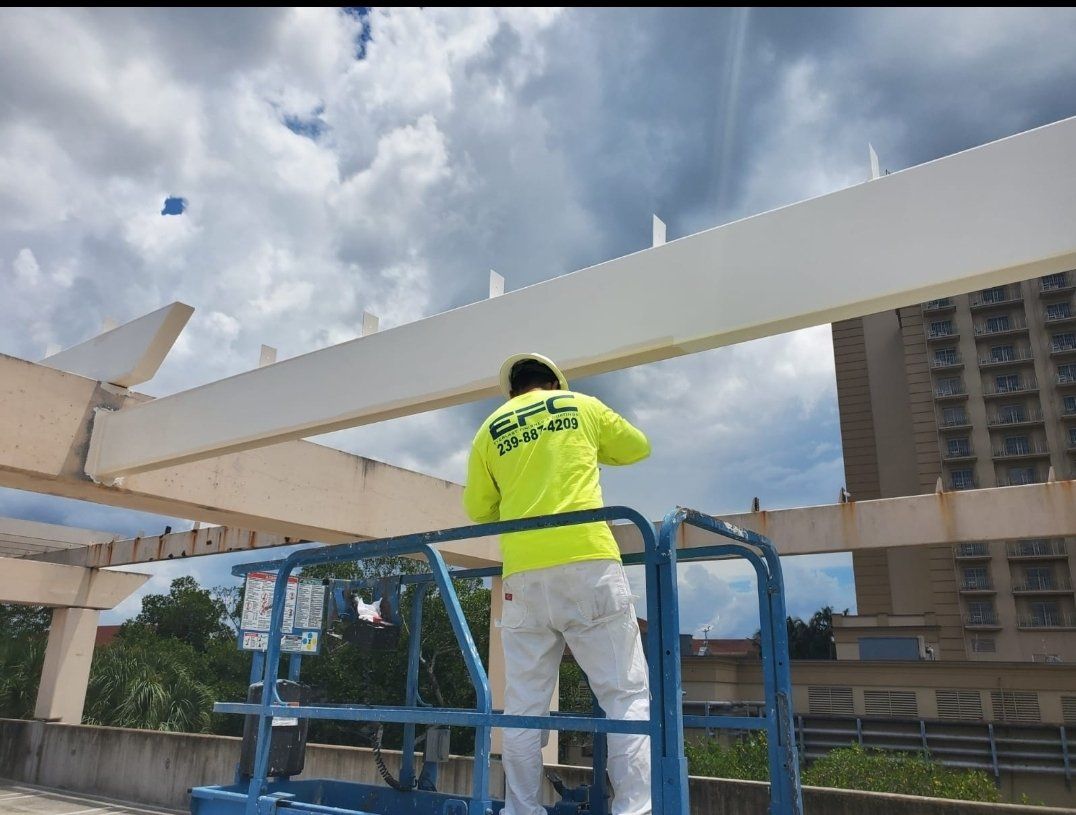 Man in neon shirt painting a white pergola from a lift under a cloudy sky.