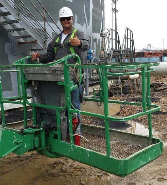 Man in lift, thumbs up, in front of industrial tank. He wears hard hat, safety vest. Outdoor setting.