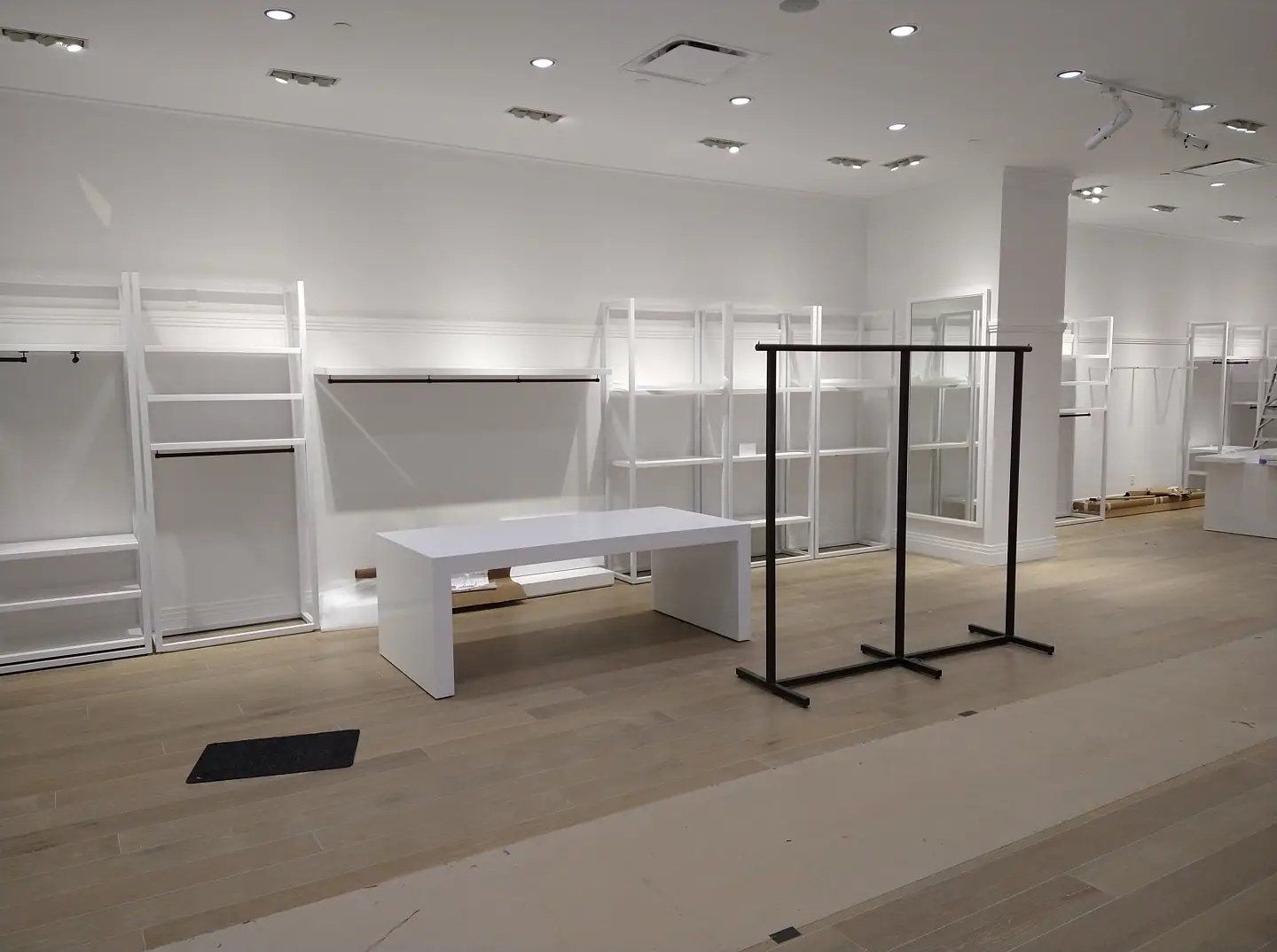 Empty retail space with white walls, shelves, and tables, plus a black clothing rack. Wooden floor.