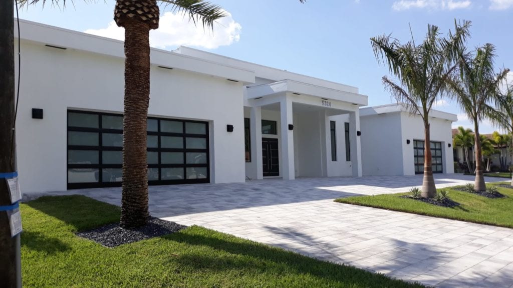 Modern white house with palm trees, glass garage doors, and a stone driveway.