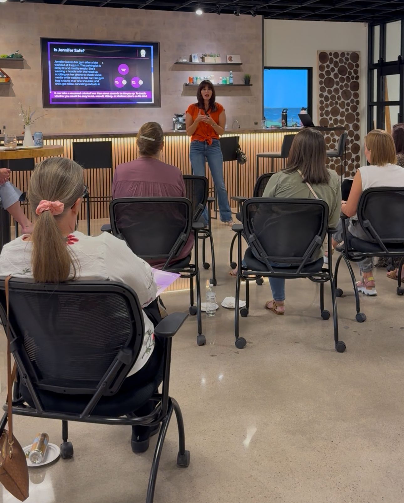Presenter speaking to seated audience in a workshop room, with a large screen displaying slides.