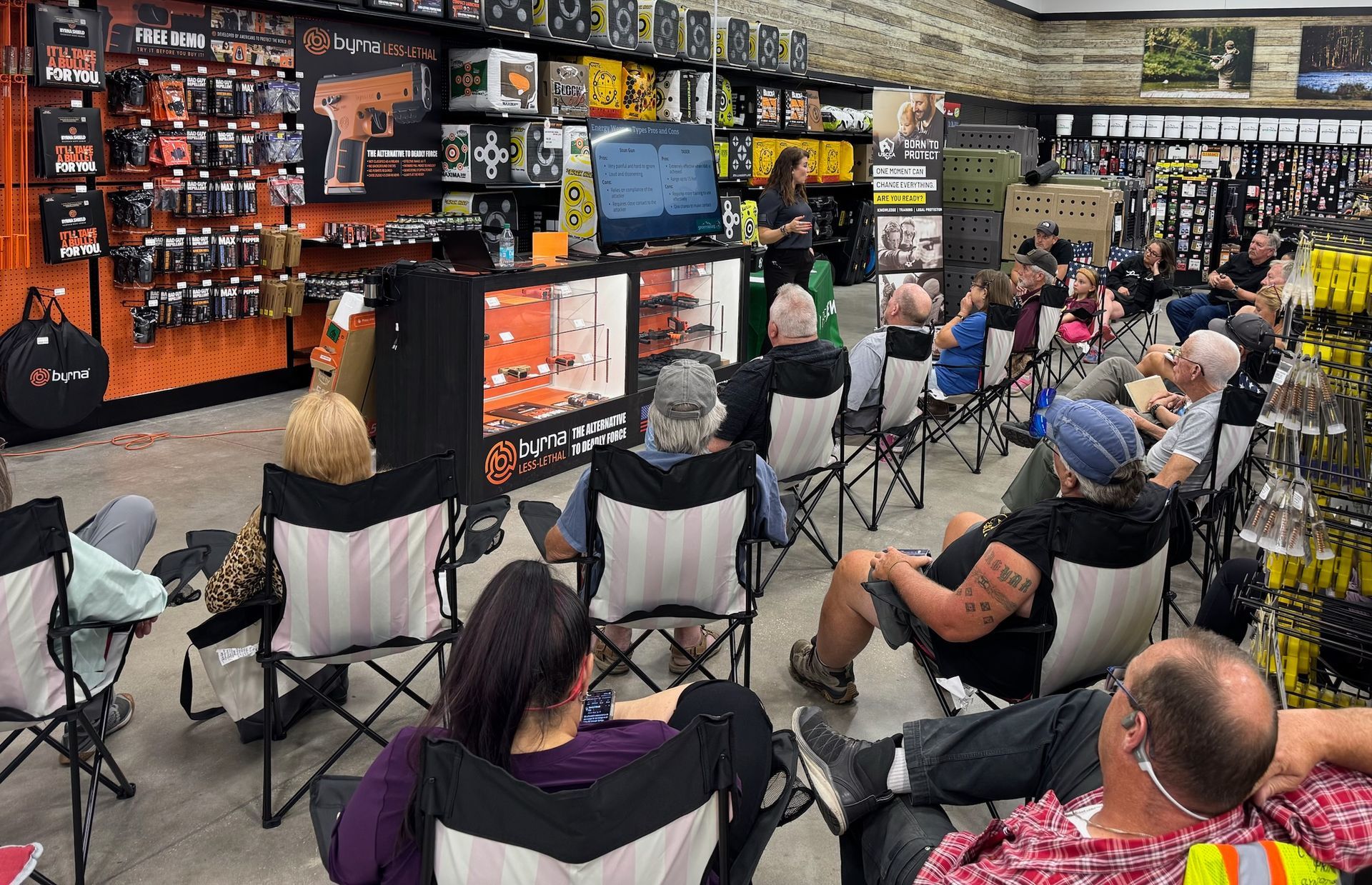 A seated audience watches a presentation in an electronics store with shelves of gear and a TV screen.
