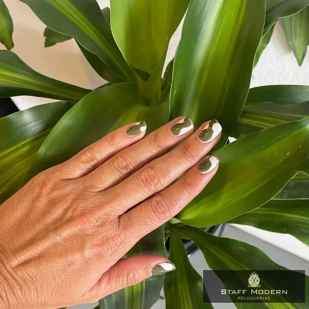 Mano con uñas de color verde metálico descansando sobre hojas de plantas verdes brillantes.