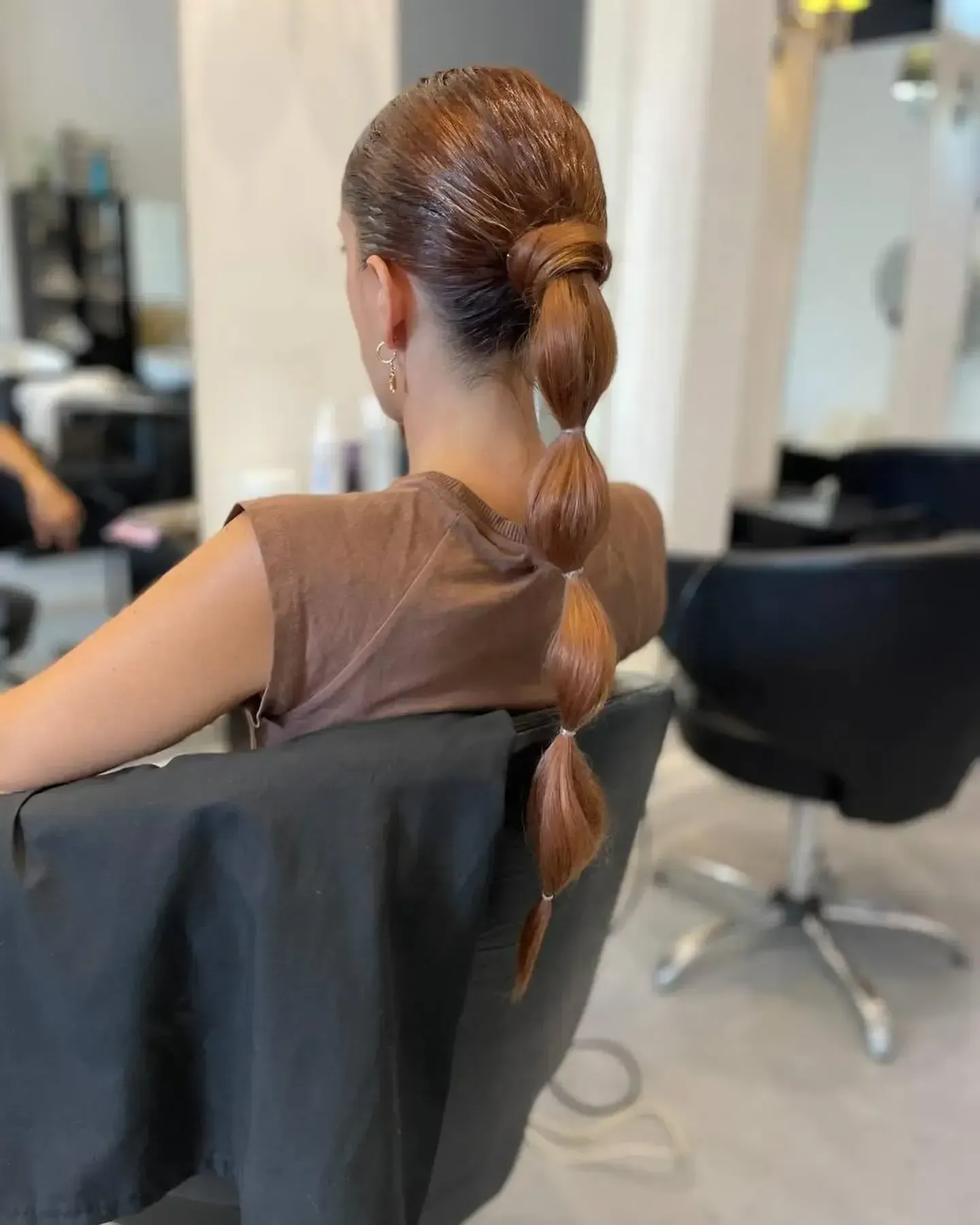Mujer con una elegante coleta trenzada sentada en una silla de peluquería, vista desde atrás.