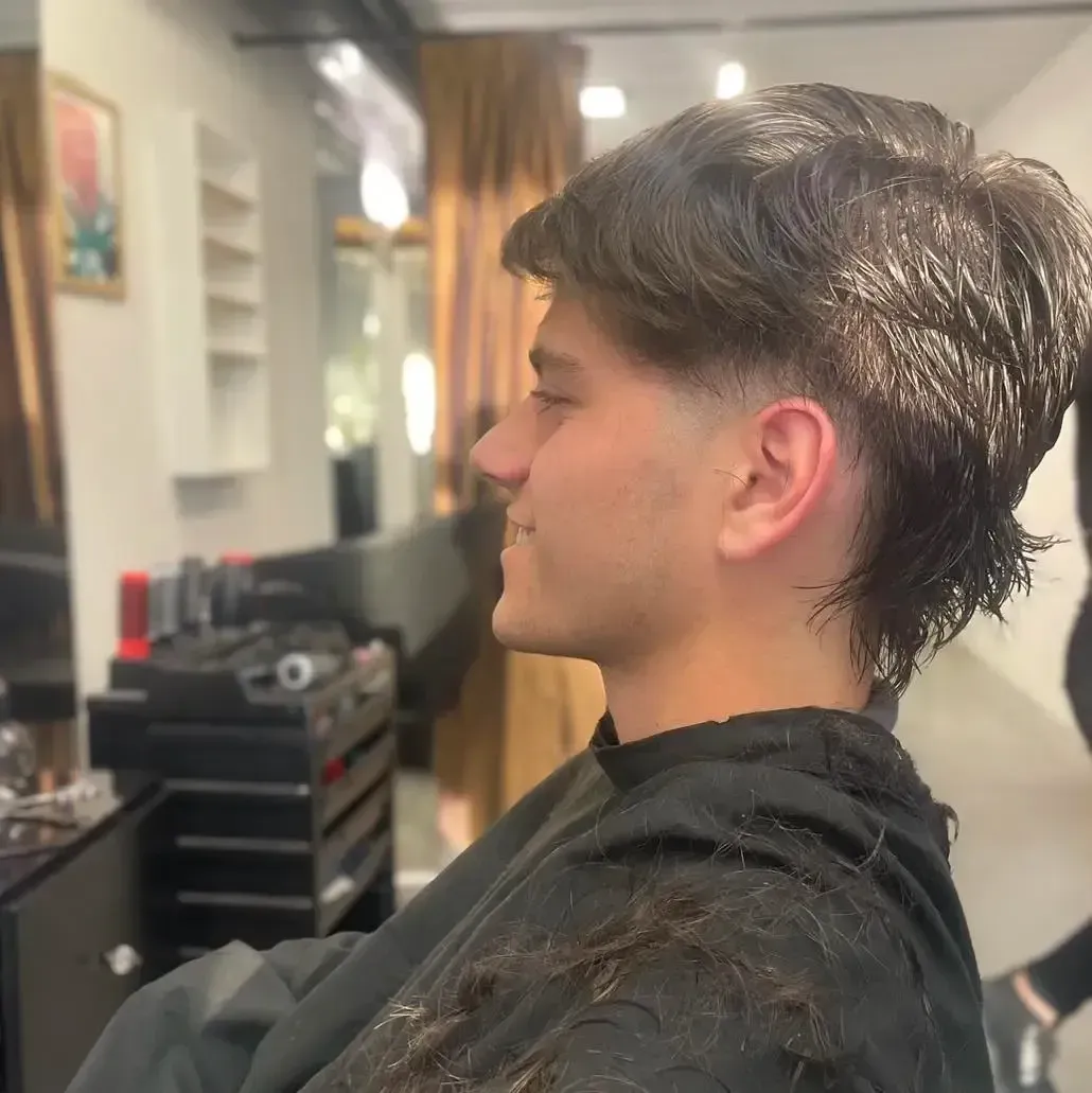 Persona de cabello oscuro sentada en una silla de peluquería durante un corte de pelo; se ve su perfil lateral.