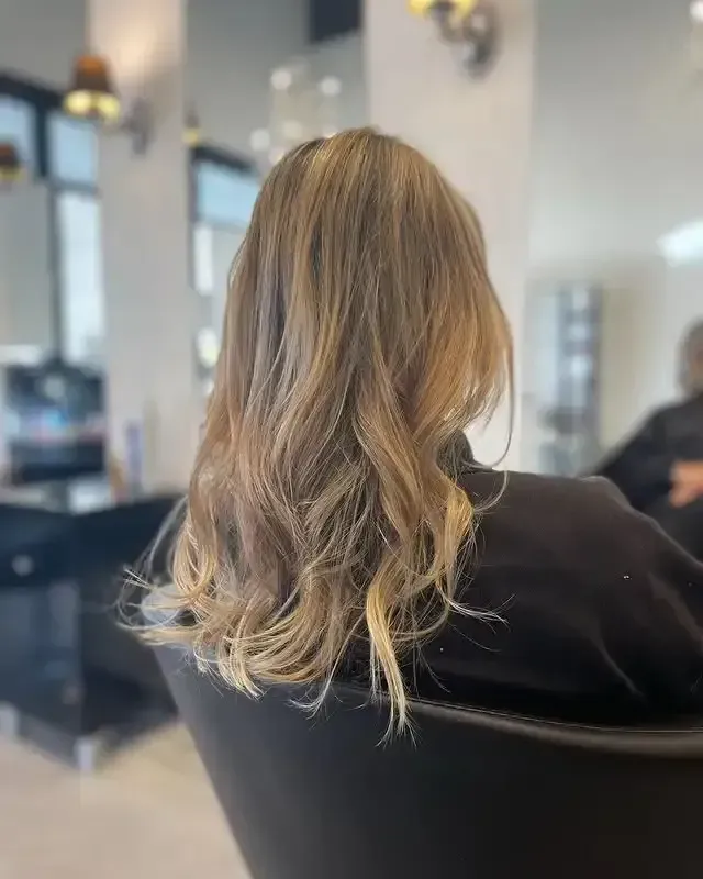 Persona con cabello rubio ondulado sentada en una silla de peluquería, vista desde atrás.