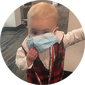Toddler in a red plaid vest holding a blue face mask to their mouth indoors