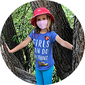 Child in red hat and pink mask standing in a tree trunk opening, wearing a blue shirt and turquoise pants.