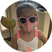 Child indoors wearing pink sunglasses and a matching face mask, holding a lemon-shaped drink cup.