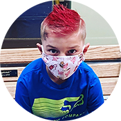 Person with bright red hair wearing a floral face mask and blue shirt, seated on a bench