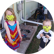 Two children in Halloween masks standing on a porch, one in a rainbow dress and one holding a treat bucket