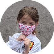 Child wearing a heart-patterned face mask and white jacket, holding the mask straps against a gray background.