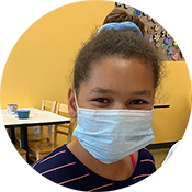 Child wearing a blue face mask in a classroom with yellow walls and tables