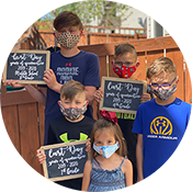 Five masked students hold small chalkboard signs outdoors in front of a brick wall.