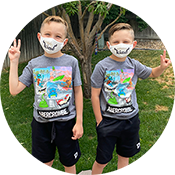 Two children in masks standing outdoors on grass, wearing matching graphic T-shirts and dark shorts