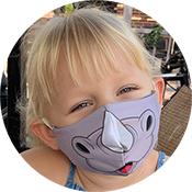 Child wearing a gray animal mask, smiling outdoors with a wicker chair in the background